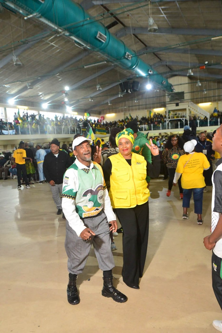 Two attendees pose on the hall floor with a gathering behind them.